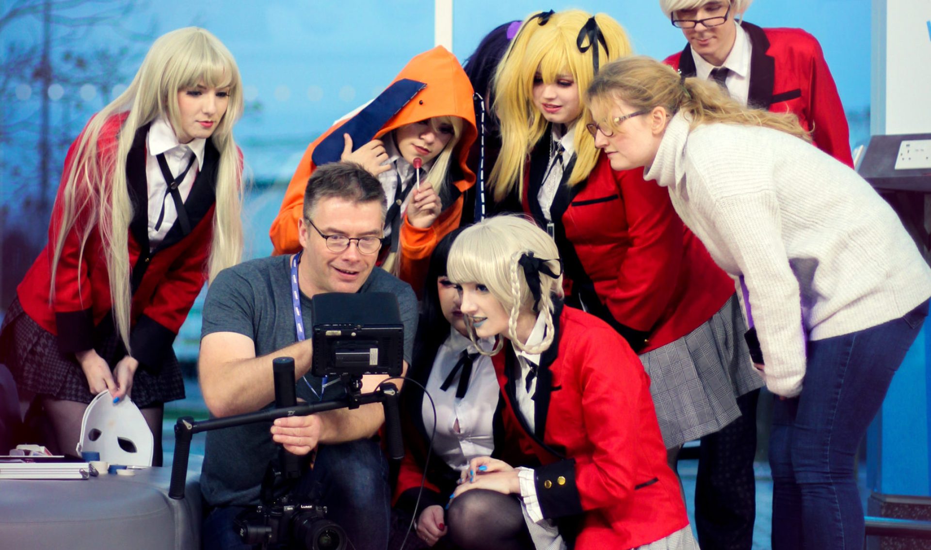 Cameraman holding a video camera on a gimbal showing what he just filmed to a group of cosplayers dressed as characters from the anime Kakegurui.